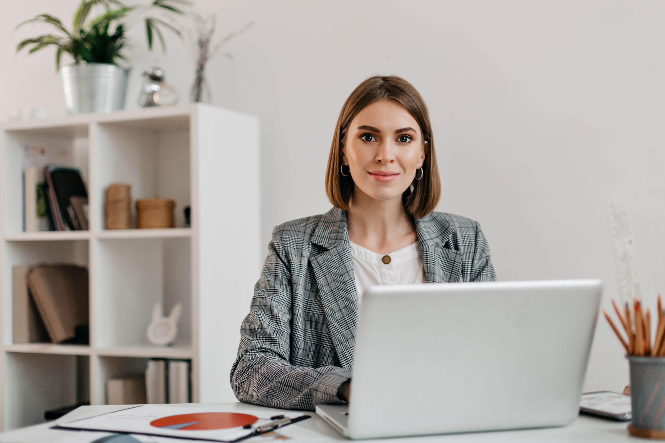 Business woman in checkered jacket looking at camera with smile while sitting at desk in her office.